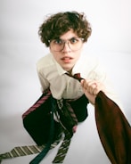 A person with curly hair and glasses is wearing a white shirt and is surrounded by numerous neckties, some wrapped around their waist, and holding a red tie in their hand. The expression appears curious or slightly bewildered.