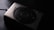 A compact digital camera with a sleek metallic design, featuring a prominent lens and branding. The lighting casts a shadow, creating a moody atmosphere.