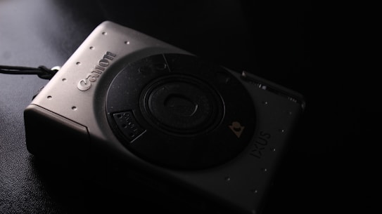 A compact digital camera with a sleek metallic design, featuring a prominent lens and branding. The lighting casts a shadow, creating a moody atmosphere.