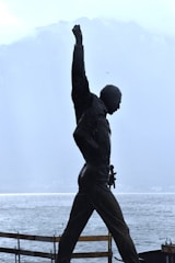 A silhouette of a statue depicting a person with one arm raised triumphantly against a backdrop of misty mountains and a body of water. The scene conveys a sense of strength and achievement, with muted colors and a serene landscape.