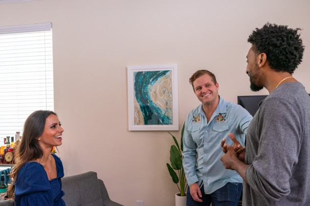 Three people are engaged in a conversation in a living room setting. A woman in a blue top is sitting and smiling at a man in a light blue shirt who is standing. Another man in a gray sweater is also part of the conversation. Behind them, there is a framed abstract artwork on the wall and a potted plant. Bottles are visible on a shelf near a window with blinds.