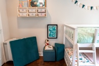 A children's room features a white bunk bed with a mint green quilt on the upper bunk. A colorful, patterned mattress is visible on the lower bunk. Teal cushions and a small teal ottoman sit nearby, with a stuffed toy on top. Wall shelves hold storage bins and children's books or drawings. A framed abstract painting hangs below the shelves. A festive banner with teal and white flags decorates the wall.