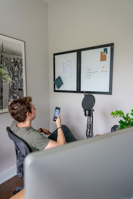 A person seated in an office chair holding a smartphone, facing a wall with two whiteboards. The whiteboards contain some written notes and papers attached with magnets. A microphone setup is visible next to the whiteboards. There's a framed black and white cityscape photo on the wall and a small green plant on the table.