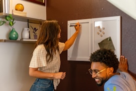 Two people are interacting with a whiteboard that is attached to a dark brown wall. A person with long hair writes on the board, while another person with glasses stands nearby, smiling. The wall features shelves with various decorative items, including books, a green vase, and a white ceramic pitcher.