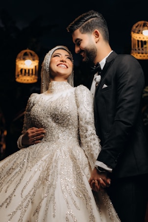 A joyful couple is dressed in wedding attire, with the bride wearing an ornate white gown embellished with intricate beadwork and the groom in a classic black suit. They are holding hands and smiling warmly at each other under soft, glowing lights that lend a romantic atmosphere.