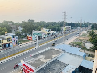 An overhead view of a busy urban street lined with buildings on one side and green spaces on the other. Various vehicles such as scooters, motorcycles, and an auto rickshaw travel along the road, which is divided by a central barrier. There are some pedestrians and a bus, as well as buildings with colorful signs.