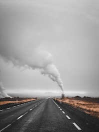 A long, empty road stretches into the distance with thick, white smoke billowing from a facility on the right side. The surrounding landscape is barren with sparse vegetation, and the sky is overcast, giving the scene a bleak atmosphere.