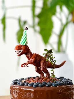 A toy dinosaur wearing a green and white striped party hat stands on top of a chocolate-covered cake. The cake is adorned with blueberries and nuts, and there is a small branch with leaves next to the dinosaur. In the background, green leaves are softly blurred.