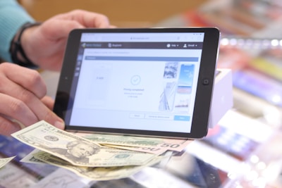 A person holding a tablet with a digital interface displaying a transaction completion message. Various denominations of U.S. currency are laid out on the counter in front of the tablet.