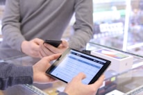 Two people are interacting over a glass counter display. One person is holding a smartphone while the other is holding a tablet displaying some sort of data or spreadsheet. There is a box for a phone or electronic device on the counter.