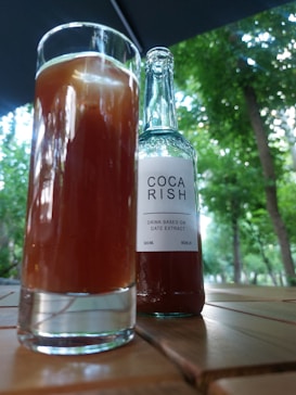 A glass filled with a brown liquid is placed on a wooden table next to a bottle labeled 'Coca Rish,' which is described as a drink based on date extract. The background displays green trees and an outdoor setting, suggesting a relaxed, natural environment.