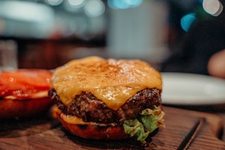 A close-up of a cheeseburger with a juicy beef patty topped with melted cheese on a toasted bun. There are slices of tomato and green lettuce visible underneath the patty, and the background is blurred, suggesting it's taken in a restaurant setting.
