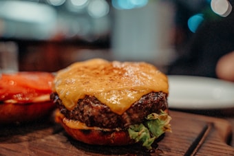 A close-up of a cheeseburger with a juicy beef patty topped with melted cheese on a toasted bun. There are slices of tomato and green lettuce visible underneath the patty, and the background is blurred, suggesting it's taken in a restaurant setting.