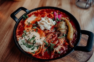 A cooked dish in a black skillet featuring vibrant red sauce, poached eggs, garnished with parsley and other spices. There are visible whole chili peppers and dollops of creamy white sauce. The wooden table beneath enhances the rustic presentation.