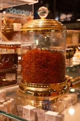 A glass jar with a gold-colored lid filled with a large quantity of saffron strands. The jar is elegantly designed with decorative elements and a crystal-like handle on top. It is placed on a glass shelf surrounded by other spice containers and small boxes.