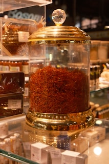A glass jar with a gold-colored lid filled with a large quantity of saffron strands. The jar is elegantly designed with decorative elements and a crystal-like handle on top. It is placed on a glass shelf surrounded by other spice containers and small boxes.