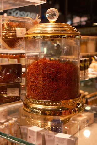 A glass jar with a gold-colored lid filled with a large quantity of saffron strands. The jar is elegantly designed with decorative elements and a crystal-like handle on top. It is placed on a glass shelf surrounded by other spice containers and small boxes.