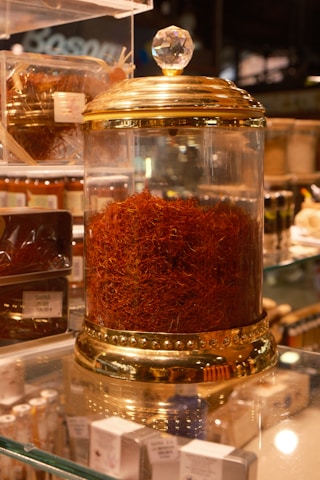 A glass jar with a gold-colored lid filled with a large quantity of saffron strands. The jar is elegantly designed with decorative elements and a crystal-like handle on top. It is placed on a glass shelf surrounded by other spice containers and small boxes.