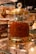 A glass jar with a gold-colored lid filled with a large quantity of saffron strands. The jar is elegantly designed with decorative elements and a crystal-like handle on top. It is placed on a glass shelf surrounded by other spice containers and small boxes.