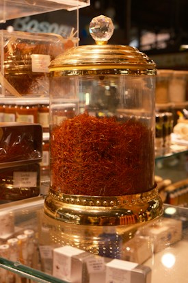 A glass jar with a gold-colored lid filled with a large quantity of saffron strands. The jar is elegantly designed with decorative elements and a crystal-like handle on top. It is placed on a glass shelf surrounded by other spice containers and small boxes.