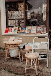 A cozy, street-side bookstall is set up outside a shop with a sign that reads 'Antykwariat', meaning an antiquarian bookstore. Various books are displayed on the window ledge and on rattan tables in front. The books range in subjects from history and art to guides about Gdansk. A couple of rattan stools are placed nearby, and the pavement is made of square tiles.