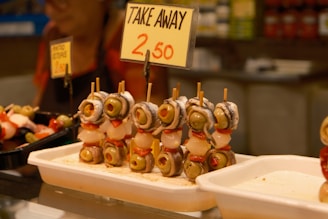 A display of skewered snacks featuring olives, peppers, onions, and anchovies is arranged in a market setting. A sign above the skewers indicates a takeaway price of 2.50. The surrounding area includes trays with additional food items and a vaguely visible person in the background.