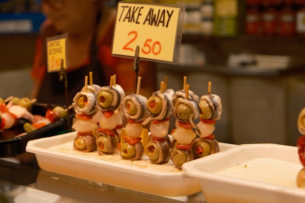A display of skewered snacks featuring olives, peppers, onions, and anchovies is arranged in a market setting. A sign above the skewers indicates a takeaway price of 2.50. The surrounding area includes trays with additional food items and a vaguely visible person in the background.