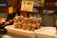 A display of skewered snacks featuring olives, peppers, onions, and anchovies is arranged in a market setting. A sign above the skewers indicates a takeaway price of 2.50. The surrounding area includes trays with additional food items and a vaguely visible person in the background.