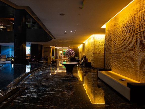 A luxurious hotel lobby features dark polished marble floors with reflections that enhance the depth of the space. Soft yellow lighting emanates from the walls, providing a warm, inviting atmosphere. In the center is a round table adorned with a large bouquet of pink flowers, adding a touch of elegance. Comfortable seating lines the walls, providing resting areas for guests. The modern aesthetic is complemented by the sleek design and minimalist decor.