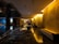 A luxurious hotel lobby features dark polished marble floors with reflections that enhance the depth of the space. Soft yellow lighting emanates from the walls, providing a warm, inviting atmosphere. In the center is a round table adorned with a large bouquet of pink flowers, adding a touch of elegance. Comfortable seating lines the walls, providing resting areas for guests. The modern aesthetic is complemented by the sleek design and minimalist decor.