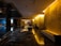 A luxurious hotel lobby features dark polished marble floors with reflections that enhance the depth of the space. Soft yellow lighting emanates from the walls, providing a warm, inviting atmosphere. In the center is a round table adorned with a large bouquet of pink flowers, adding a touch of elegance. Comfortable seating lines the walls, providing resting areas for guests. The modern aesthetic is complemented by the sleek design and minimalist decor.