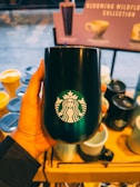 A hand holding a black tumbler with the Starbucks logo, surrounded by various other cups and tumblers displayed on a wooden shelf. In the background, there's a blurred sign with text indicating a collection of drinkware.