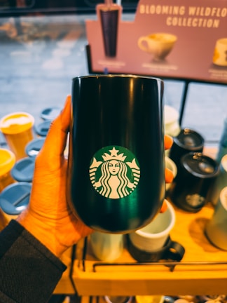A hand holding a black tumbler with the Starbucks logo, surrounded by various other cups and tumblers displayed on a wooden shelf. In the background, there's a blurred sign with text indicating a collection of drinkware.