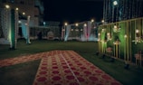 An elaborately decorated outdoor space set up for an event, possibly a wedding or celebration. The area is adorned with strings of lights, floral arrangements, and draped fabric. Red and gold carpets are laid over the grass, leading to a central spot with a backdrop for photos or seating. Warm lighting creates an inviting and festive atmosphere.