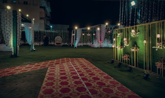 An elaborately decorated outdoor space set up for an event, possibly a wedding or celebration. The area is adorned with strings of lights, floral arrangements, and draped fabric. Red and gold carpets are laid over the grass, leading to a central spot with a backdrop for photos or seating. Warm lighting creates an inviting and festive atmosphere.