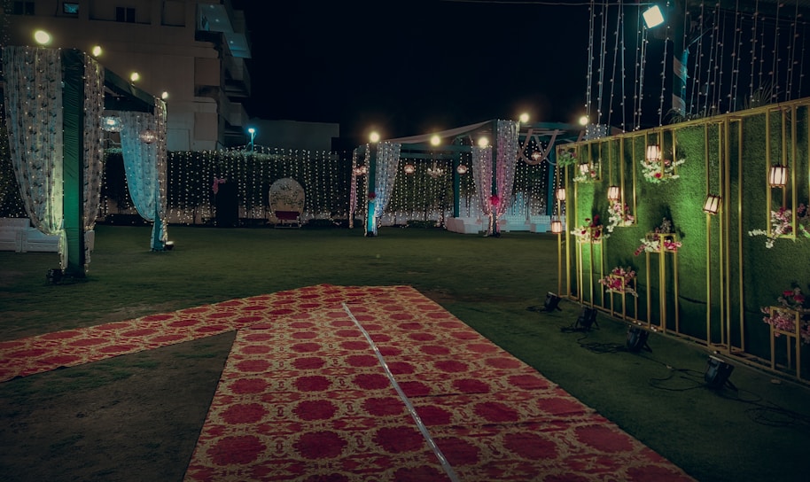 An elaborately decorated outdoor space set up for an event, possibly a wedding or celebration. The area is adorned with strings of lights, floral arrangements, and draped fabric. Red and gold carpets are laid over the grass, leading to a central spot with a backdrop for photos or seating. Warm lighting creates an inviting and festive atmosphere.