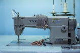 A heavy-duty industrial sewing machine is placed on a table, with a large spool of white thread positioned in the background. Scissors and a few crafting tools are laid out on the table next to the machine. There is a caution sign on the machine, indicating safety instructions. The background features a light blue wall, contributing to a clean and organized workspace appearance.