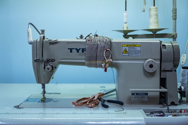 A heavy-duty industrial sewing machine is placed on a table, with a large spool of white thread positioned in the background. Scissors and a few crafting tools are laid out on the table next to the machine. There is a caution sign on the machine, indicating safety instructions. The background features a light blue wall, contributing to a clean and organized workspace appearance.