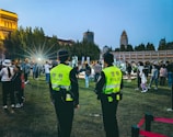 Officers in bright yellow vests are overseeing a gathering in an outdoor area during the evening. The crowd includes people of various ages and the background is composed of illuminated historical architecture with a city skyline featuring modern skyscrapers.
