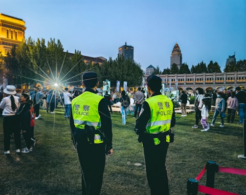 Officers in bright yellow vests are overseeing a gathering in an outdoor area during the evening. The crowd includes people of various ages and the background is composed of illuminated historical architecture with a city skyline featuring modern skyscrapers.