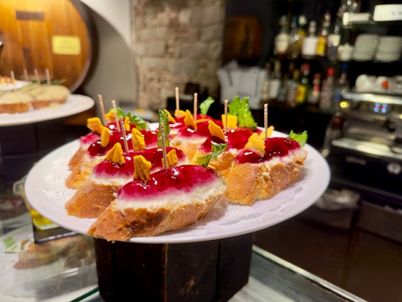 A white plate holds several slices of bread topped with a layer of creamy cheese and a glossy red sauce. Garnished with vibrant green mint leaves and small yellow chips, each piece is skewered with a toothpick for easy handling. The background features a rustic setting with bottles arranged on shelves, adding a cozy atmosphere.