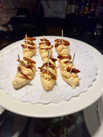 Appetizers displayed on a white plate with a delicate doily underneath. The appetizers consist of small pieces of bread topped with creamy spread and sun-dried tomatoes, each secured with a toothpick.