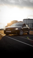 A sleek black Audi car is parked on a pavement with the sun setting behind tall modern buildings. The car's headlights are on, and the front license plate is visible. The image captures a blend of urban architecture and an automotive focus.