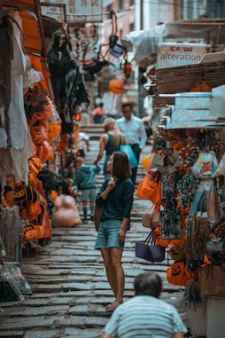 A bustling market street with various Halloween decorations and merchandise hanging from stalls. People are casually walking and browsing the items on display. The decorations mainly consist of orange pumpkin-themed items and spooky figures. Stone steps create a narrow pathway through the market.