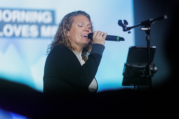 A person with curly hair sings passionately into a microphone on stage. The background features a softly blurred screen with partially visible text. The person is wearing a dark jacket over a light-colored shirt, and there is a stand with a tablet or device in front of them.