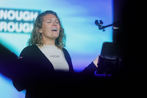 A person is singing passionately with eyes closed, holding a microphone. The background features a blurred blue and white screen with text partially visible.
