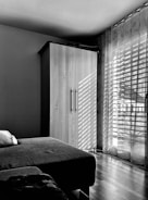 A minimalist bedroom interior with a wooden wardrobe and a bed with a pillow. Sunlight filters through sheer curtains, casting striped shadows on the wardrobe and wooden floor.