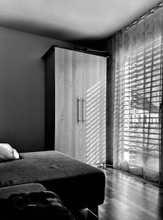 A minimalist bedroom interior with a wooden wardrobe and a bed with a pillow. Sunlight filters through sheer curtains, casting striped shadows on the wardrobe and wooden floor.