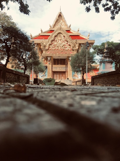 A grand, ornate temple with intricate carvings and golden details dominates the image. The temple features a thick red roof, decorative elements, statues, and a symmetrical facade. It is surrounded by trees and greenery, and the ground leading up to it is covered in leaves and textured stones. The sky is partly cloudy, adding depth to the scene.