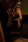 Two hands reaching towards each other in a dimly lit environment, with a focus on the expressive gesture of the fingers. The setting suggests a moment of connection or support, with clothing indicating casual attire.